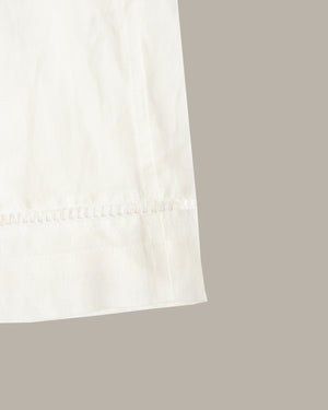 Close-up of a white fabric product corner showing a folded hem with a subtle scalloped or ruffled edge, set against a plain warm gray background.