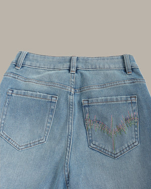 Close-up back view of light blue denim jeans showing the waistband, belt loops, and back pockets, with multicolored embroidered details on the right pocket.