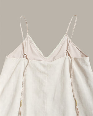 A close-up of a light cream or off-white camisole top with thin braided spaghetti straps, a V-neckline with delicate lace trim, and a relaxed draped silhouette, shown against a solid warm gray background.