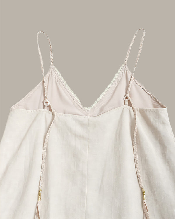 A close-up of a light cream or off-white camisole top with thin braided spaghetti straps, a V-neckline with delicate lace trim, and a relaxed draped silhouette, shown against a solid warm gray background.