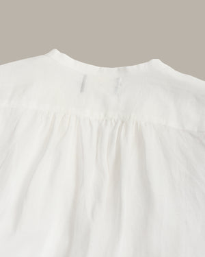 Close-up of a white textured fabric garment, showing a gathered neckline and fine weave, set against a solid taupe background.
