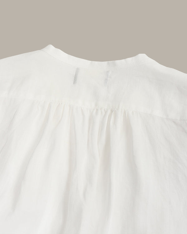 Close-up of a white textured fabric garment, showing a gathered neckline and fine weave, set against a solid taupe background.