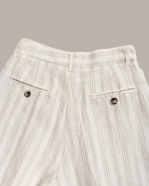 Close-up of a pair of off-white linen trousers with vertical brown stripes, featuring two buttoned flap pockets and a pleated waistband, set against a solid taupe background.