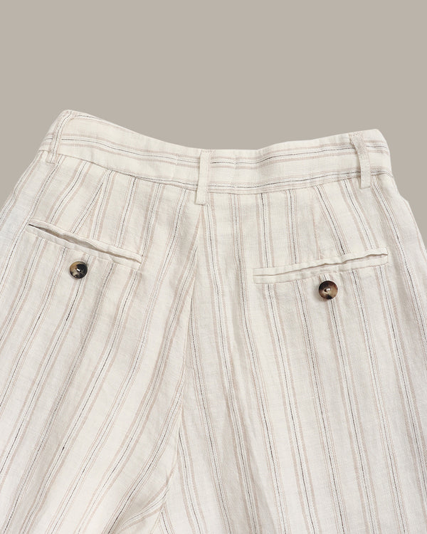 Close-up of a pair of off-white linen trousers with vertical brown stripes, featuring two buttoned flap pockets and a pleated waistband, set against a solid taupe background.