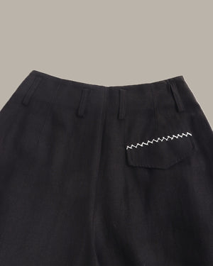 Close-up of black trousers showing the waistband, front fly, and a side pocket with distinctive zigzag white stitching, set against a solid taupe background.