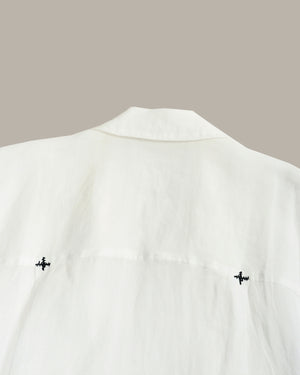 Close-up of a white shirt or blouse showing the back collar and upper back, with two small black embroidered motifs near the lower sides, against a plain warm gray background.