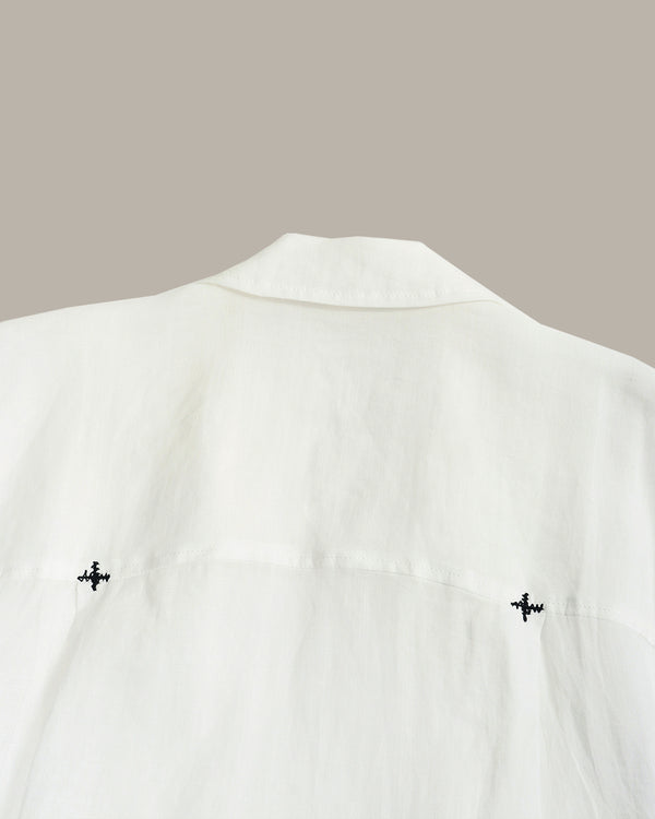 Close-up of a white shirt or blouse showing the back collar and upper back, with two small black embroidered motifs near the lower sides, against a plain warm gray background.
