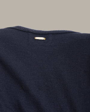 Close-up of the back of a dark navy knit sweater, showing a small white fabric brand tag with gold lettering near the neckline, set against a solid taupe background.