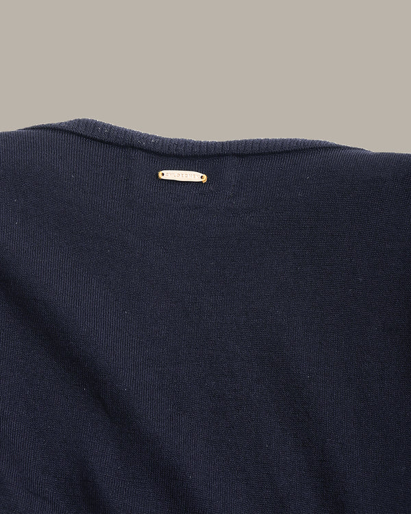 Close-up of the back of a dark navy knit sweater, showing a small white fabric brand tag with gold lettering near the neckline, set against a solid taupe background.