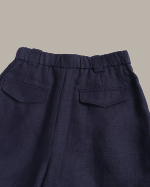 Close-up of the back waistband and pockets of dark navy blue trousers, showing a pleated elastic waistband and two flap pockets against a solid taupe background.
