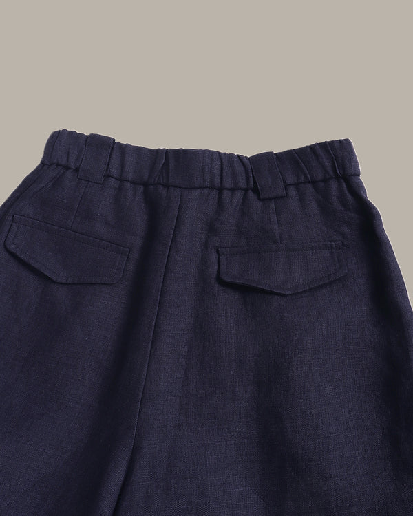 Close-up of the back waistband and pockets of dark navy blue trousers, showing a pleated elastic waistband and two flap pockets against a solid taupe background.