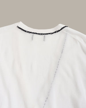 Close-up of a white top or dress neckline with a black chain detail along the collar and a vertical chain seam running down the front, shown against a plain taupe background.