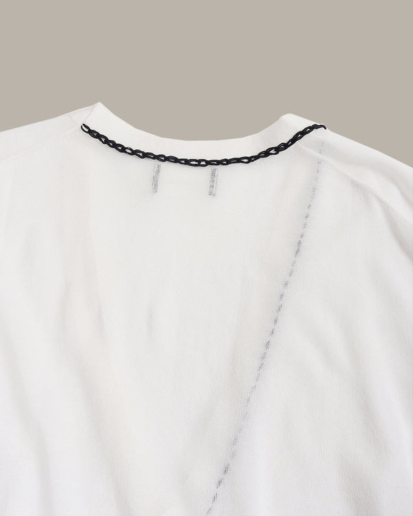 Close-up of a white top or dress neckline with a black chain detail along the collar and a vertical chain seam running down the front, shown against a plain taupe background.