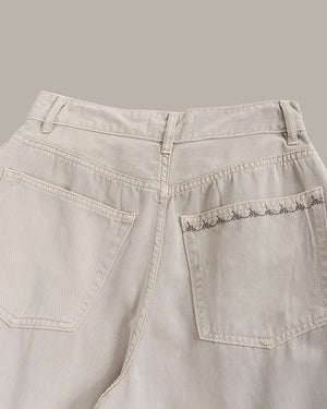 Close-up of the back waistband and rear pockets of light beige denim jeans, showing a decorative scalloped embroidery on the right pocket.