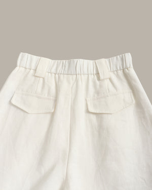 Close-up of the waistband and back pockets of off-white linen trousers, showing a gathered elastic waist and two welt pockets, set against a solid taupe background.