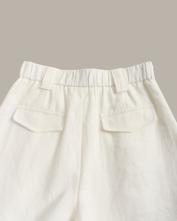 Close-up of the waistband and back pockets of off-white linen trousers, showing a gathered elastic waist and two welt pockets, set against a solid taupe background.