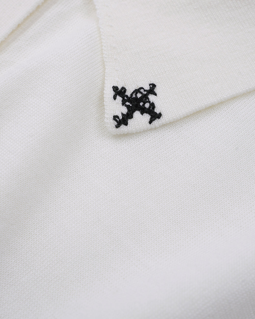 A white long-sleeve button-up shirt with a pointed collar, featuring small black embroidered star details on either side of the collar and light-colored buttons, displayed against a solid taupe background.