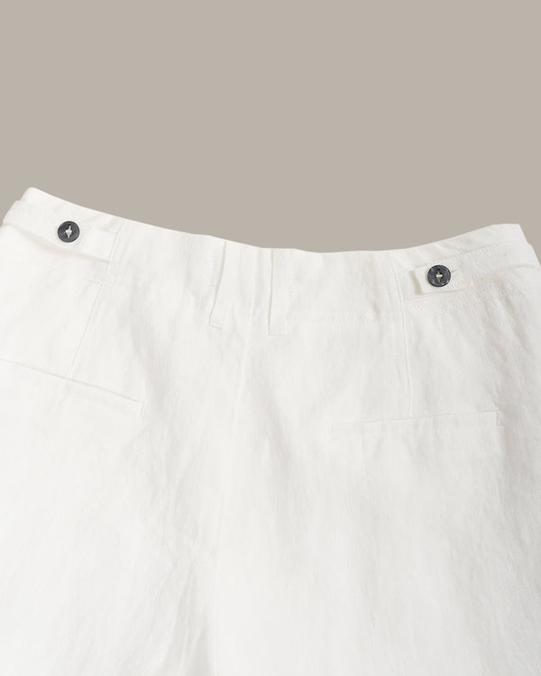 Close-up of white trousers showing the waistband with two dark buttons and front pocket details, photographed against a plain taupe background.