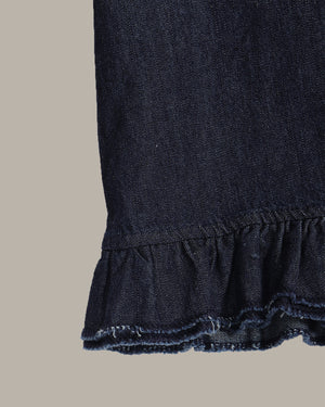 Close-up detail of a dark indigo denim garment, showing a gathered waistband and a ruffled hem with frayed edges, set against a solid light gray background.