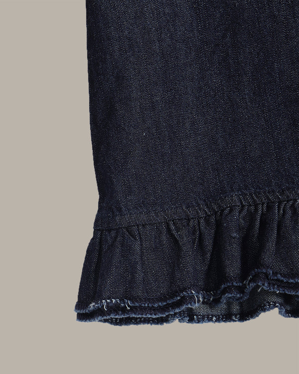 Close-up detail of a dark indigo denim garment, showing a gathered waistband and a ruffled hem with frayed edges, set against a solid light gray background.
