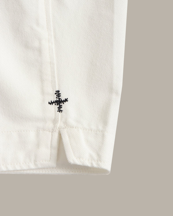 Close-up of a white fabric garment corner with a small black embroidered cross logo and hem stitching, set against a solid taupe background.