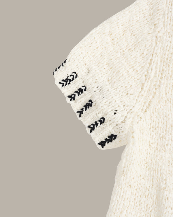 Close-up of a cream-colored knit garment sleeve with black embroidered chevron details along the cuff, set against a solid taupe background.