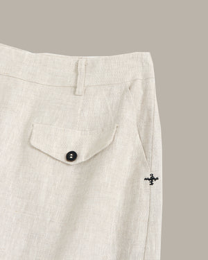 Close-up of light beige linen trousers showing a buttoned flap pocket and a small black embroidered cross logo on the side seam.