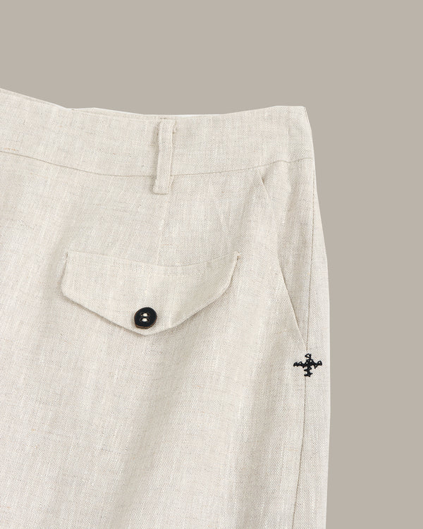 Close-up of light beige linen trousers showing a buttoned flap pocket and a small black embroidered cross logo on the side seam.