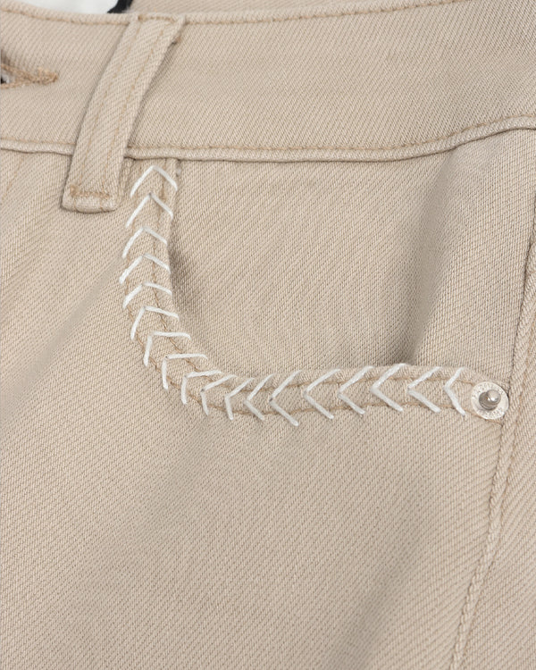 Close-up of a beige fabric garment, likely shorts or pants, showing a decorative white zigzag stitch detail near the pocket area.