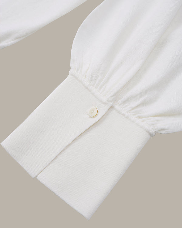 Close-up of a white long-sleeve blouse cuff with a single button closure and gathered elastic seam, shown against a plain warm beige background.