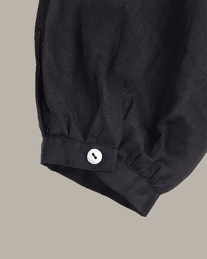 Close-up of a black fabric garment sleeve with a gathered cuff and a single white button, shown against a plain warm gray background.