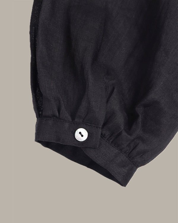 Close-up of a black fabric garment sleeve with a gathered cuff and a single white button, shown against a plain warm gray background.