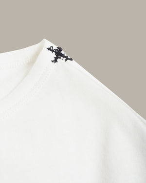 Close-up of a white fabric garment corner with a small black embroidered or printed decorative motif near the neckline, set against a plain warm gray background.