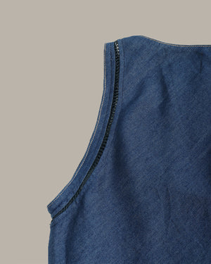 Close-up of a blue denim sleeveless top showing the shoulder seam and black contrast stitching against a solid beige background.