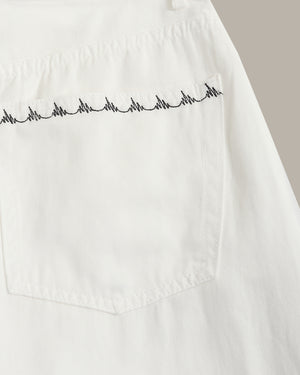 Close-up of a white fabric garment showing a pocket with black zigzag embroidery across the top edge, photographed against a light neutral background.