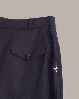Close-up of dark navy trousers showing the waistband, a buttoned flap pocket, and a small white embroidered compass logo on the side, set against a solid taupe background.