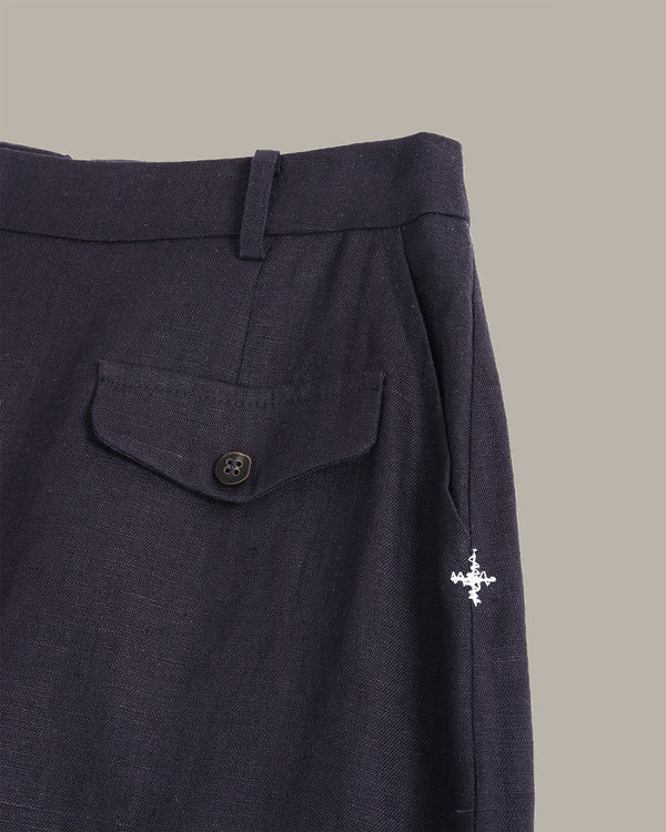 Close-up of dark navy trousers showing the waistband, a buttoned flap pocket, and a small white embroidered compass logo on the side, set against a solid taupe background.