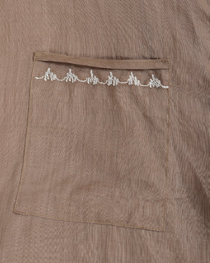 Close-up of a taupe-brown textured fabric garment featuring a front pocket with a decorative row of white embroidered details along the top edge.