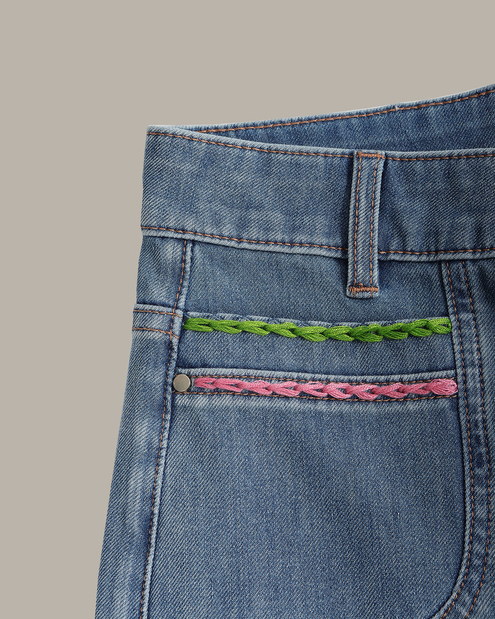 Light blue wide-leg denim jeans with colorful embroidered details on the front pockets, displayed flat against a solid taupe background.