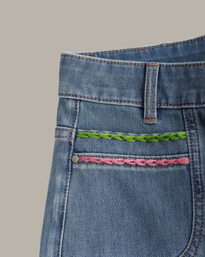 Close-up of the back pocket of blue denim jeans featuring two decorative braided embroidery accents, one green and one pink, on a solid beige background.