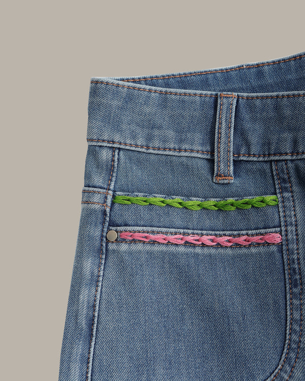 Close-up of the back pocket of blue denim jeans featuring two decorative braided embroidery accents, one green and one pink, on a solid beige background.