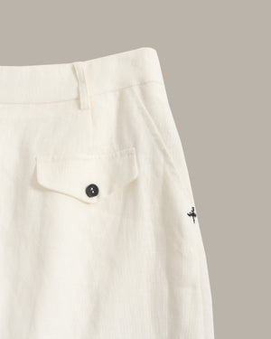 Close-up of off-white trousers showing a buttoned flap pocket, waistband, and a small black embroidered logo on the side, set against a solid taupe background.