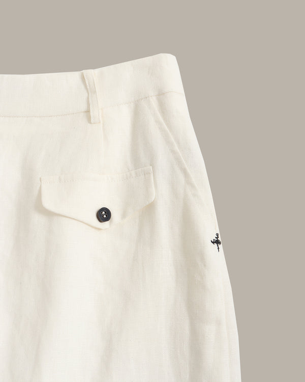 Close-up of off-white trousers showing a buttoned flap pocket, waistband, and a small black embroidered logo on the side, set against a solid taupe background.