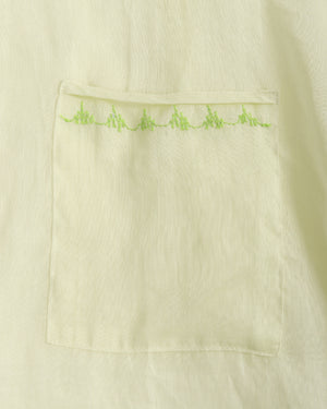 Close-up of a light cream or off-white fabric garment with a rectangular patch pocket featuring a row of small lime green embroidered details along the top edge.
