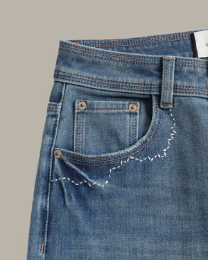 Close-up of a pair of blue denim jeans showing the back pocket with decorative white zigzag stitching, metal rivets, and contrast stitching on a solid taupe background.