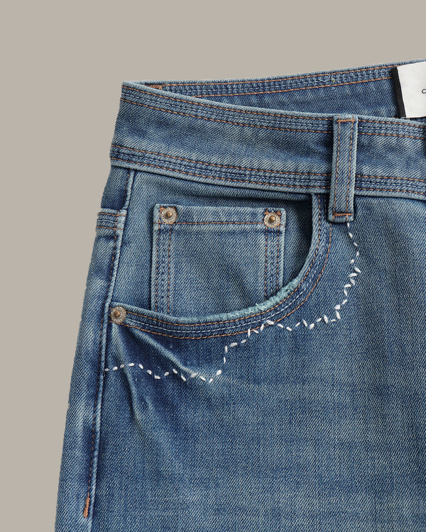 Close-up of a pair of blue denim jeans showing the back pocket with decorative white zigzag stitching, metal rivets, and contrast stitching on a solid taupe background.