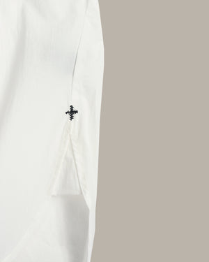 Close-up of a white garment sleeve with a small black embroidered cross detail, set against a solid taupe background.