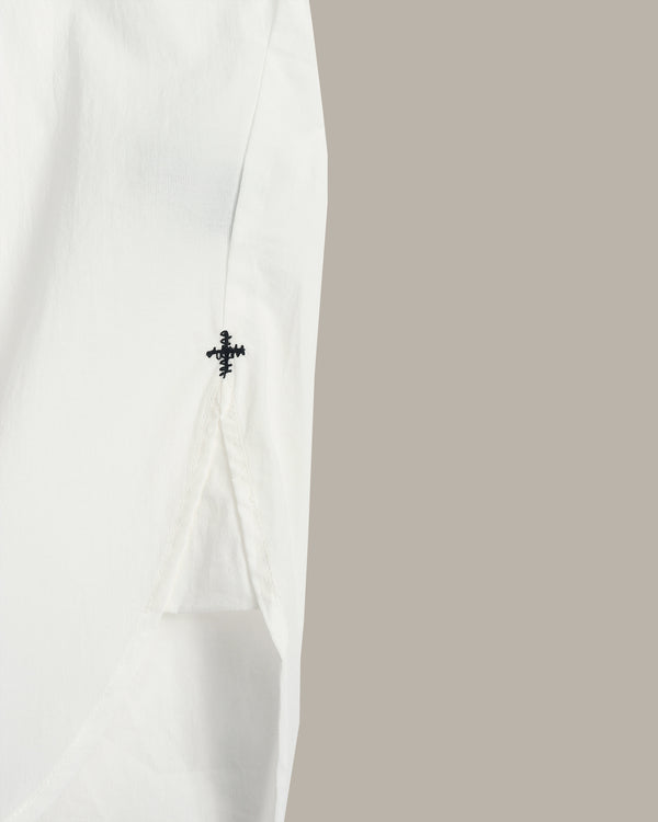 Close-up of a white garment sleeve with a small black embroidered cross detail, set against a solid taupe background.