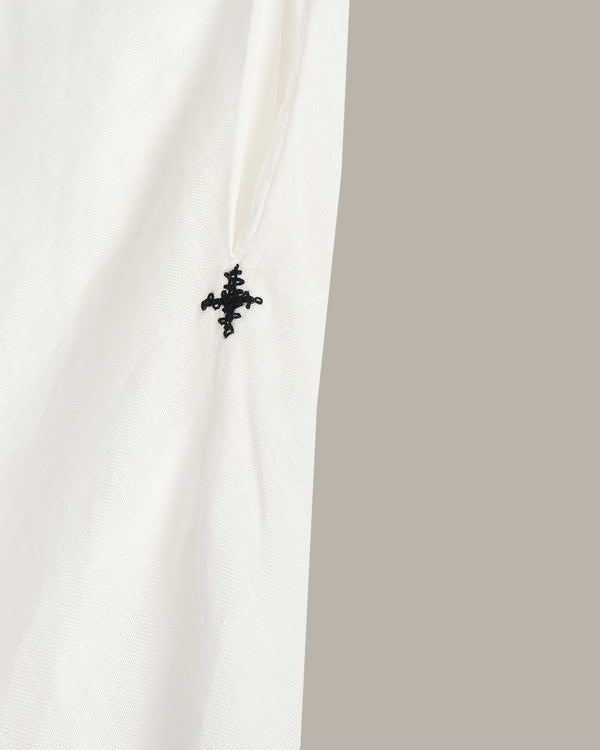 Close-up of a white textured fabric with a small black embroidered cross logo near the center, set against a solid taupe-gray background.