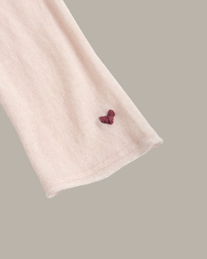 A close-up of a light pink ribbed knit fabric sleeve or cuff with a small embroidered burgundy heart near the cuff edge, set against a solid taupe background.